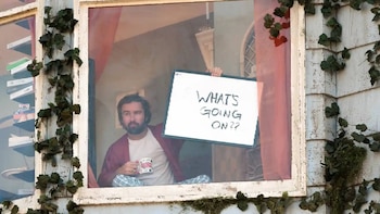 Un hombre barbudo sostiene un cartel blanco con el texto 'WHAT'S GOING ON??'. Está sentado en una ventana, con una taza en la mano. Hiedra en la pared