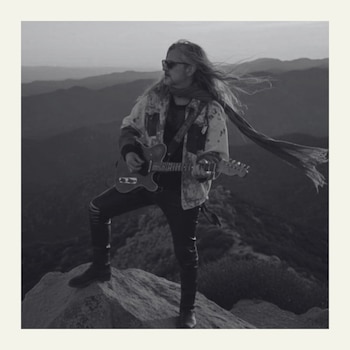 Hombre con cabello largo, gafas de sol y bufanda, toca una guitarra eléctrica sobre una roca en una montaña. Usa chaqueta, pantalones oscuros y botas