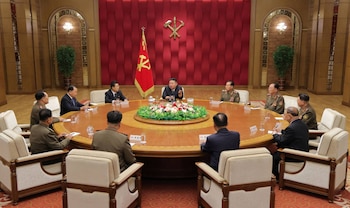 Kim Jong-un y varios hombres en trajes y uniformes militares están sentados en una gran mesa redonda ante un fondo rojo con un emblema