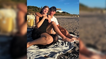 Dos jóvenes, una mujer y un hombre, se sientan en una toalla sobre la arena de una playa con conchas. Detrás se observan vegetación y un edificio blanco