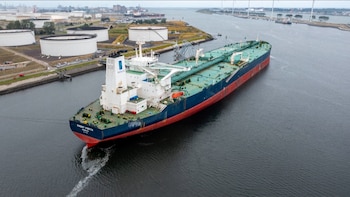 Un gran petrolero azul y rojo con superestructura blanca navega en un canal. Se aprecian tanques de almacenamiento y turbinas eólicas
