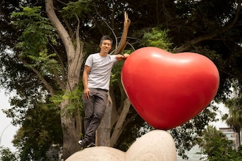 Un hombre posa apoyado en una gran escultura de un corazón rojo en un parque rodeado de árboles