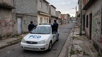 Patrulla blanca de la Policía de Control de Drogas (PCD) con dos agentes en uniforme oscuro en una calle angosta de barrio, con casas de bloques y grafitis.