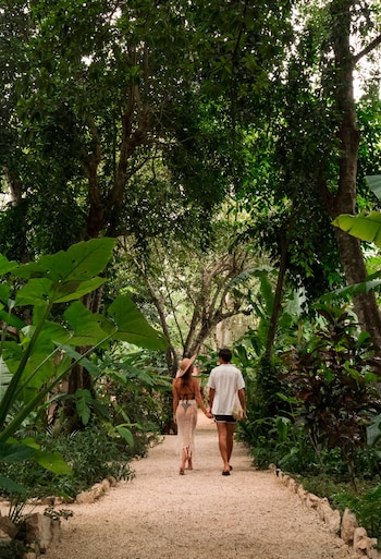 Sendero de grava con rocas en los bordes. Dos personas caminan, una mujer con sombrero y vestido largo, un hombre con camisa blanca, pantalones cortos y bolso. Vegetación densa