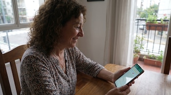 Una mujer sentada ante una mesa de madera sostiene un teléfono inteligente que exhibe una aplicación de mensajería instantánea.