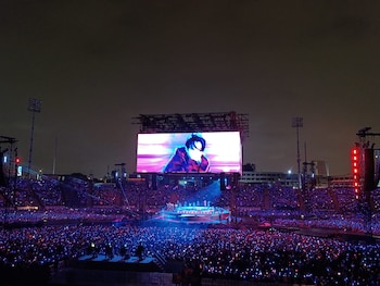 Estadio de noche repleto de personas con luces de colores, con un escenario central y una pantalla gigante que proyecta a un cantante