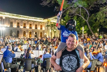 Padre lleva a hijo con máscara de Darth Vader y sable de luz rojo sobre sus hombros, sonriendo en un festival nocturno con orquesta y público de fondo