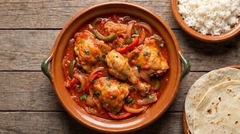 Vista aérea de un plato de barro con pollo en salsa de tomate, pimientos, cebolla, arroz blanco y tortillas sobre una mesa de madera.