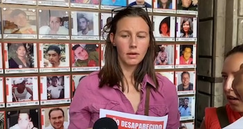 Una mujer con cabello castaño, vestida de rosa, habla frente a un muro con múltiples fotos de personas desaparecidas; sostiene un cartel que dice 'DESAPARECIDO'