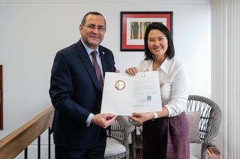 Hombre de traje y mujer con blusa blanca sostienen documento oficial con sello de Presidencia de El Salvador. Una silla y un cuadro son visibles