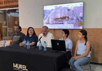 Cinco personas sentadas en una mesa con un micrófono y botellas de agua. Detrás, una pantalla exhibe rocas y texto sobre pinturas rupestres. Hay un portátil