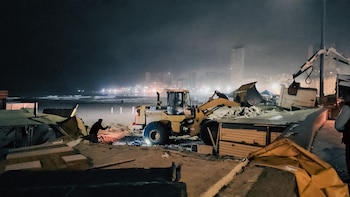 Una cargadora frontal amarilla y una excavadora blanca en una playa arenosa de noche, con una persona trabajando entre escombros y luces de la ciudad al fondo