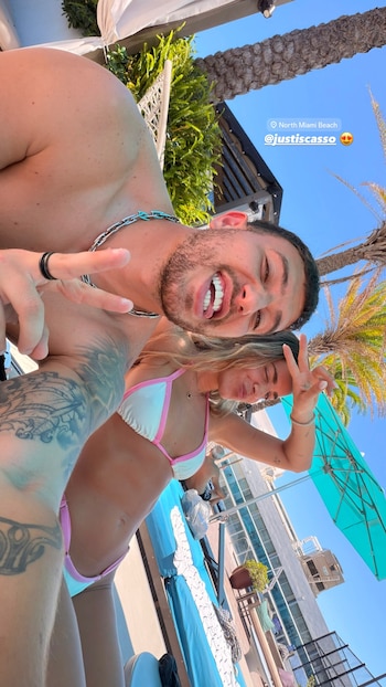 Un hombre sin camiseta y una mujer en bikini sonríen y hacen el signo de la paz en una selfie. Se ven palmeras, cielo azul y edificios. Texto: North Miami Beach