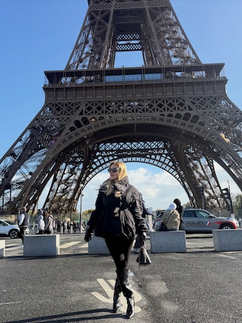 Una mujer con abrigo negro, botas y gafas oscuras camina sobre el pavimento frente a la base de la Torre Eiffel bajo un cielo despejado