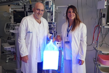 Dos médicos, un hombre y una mujer con batas blancas, posan con un dispositivo de fototerapia azul brillante en un entorno hospitalario con equipo médico
