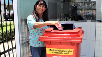 Mujer sonriente deposita una caja rosa de medicamentos en un contenedor rojo de reciclaje con letrero amarillo frente a una farmacia