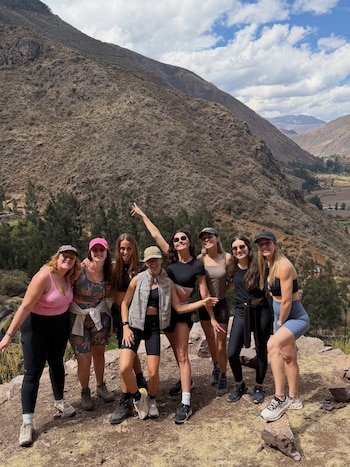 Ocho mujeres posan sonrientes en un sendero de montaña, con montañas áridas y cielo parcialmente nublado de fondo