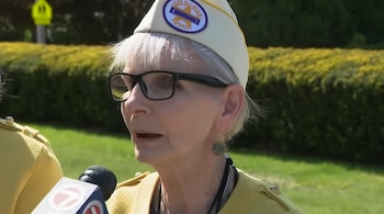 Primer plano de una mujer mayor con gafas y cabello gris, vistiendo un gorro blanco con el logo 'Gold Star Wives' y una chaqueta amarilla, hablando a un micrófono
