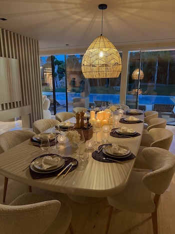 Mesa de comedor larga y blanca con 10 sillas, vajilla, copas, velas encendidas, lámpara colgante de mimbre y ventanal con vista a una piscina de noche