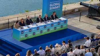 Seis personas sentadas en una mesa de conferencia al aire libre ante un público, junto al mar y con un panel promocional detrás