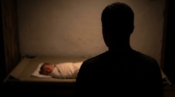 Silueta de un hombre con la espalda hacia el espectador, observando a un bebé recién nacido envuelto en una manta blanca, tumbado en una cuna en una habitación poco iluminada.