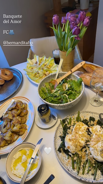 Vista aérea de una mesa blanca con varios platos de comida, incluyendo ensalada verde, papas asadas, broccolini con queso y almendras, y flores moradas en un florero