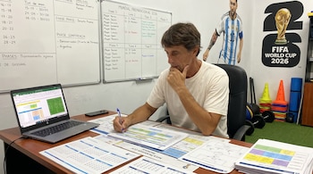 Luis Martín, preparador físico, sentado en un escritorio con un portátil y documentos, escribiendo. Detrás, una pizarra y un mural de Messi junto al logo del Mundial 2026.