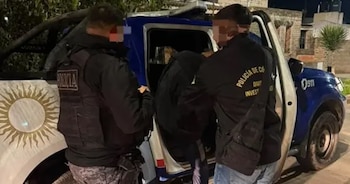 Dos agentes de policía introducen a una persona en una camioneta policial azul y blanca. Los uniformes llevan la palabra 'POLICIA' y un emblema de sol