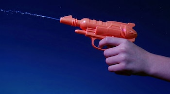 Primer plano de la mano de un adolescente sosteniendo una pistola de agua de juguete naranja que lanza un chorro de agua. Fondo azul oscuro.