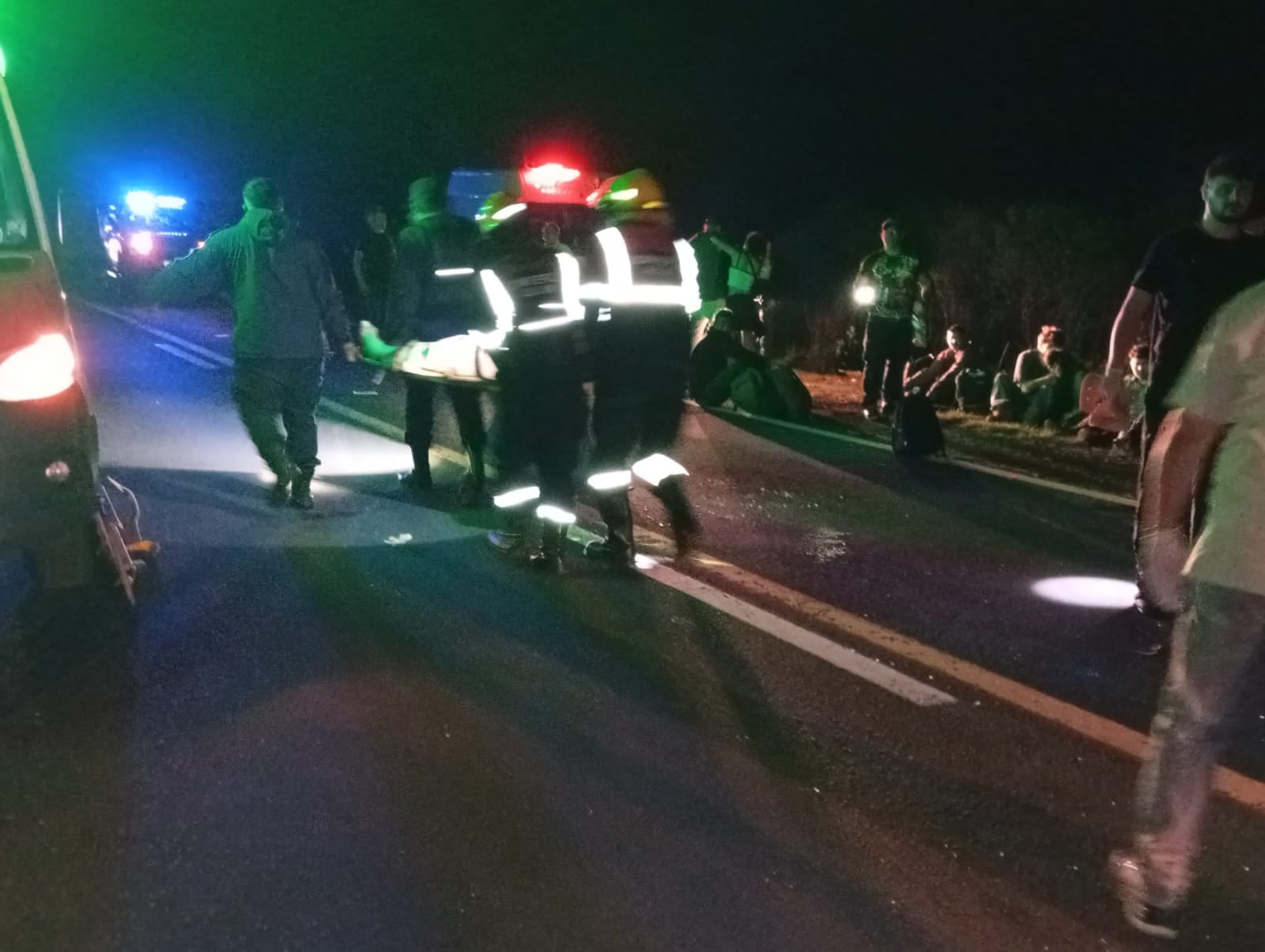 Personal de emergencia transporta a una persona en camilla, mientras el resto de los pasajeros esperan al lado de la ruta