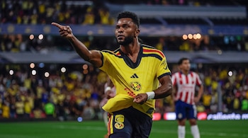 Un futbolista con uniforme amarillo señala hacia un costado con el brazo extendido en el campo de juego durante un partido nocturno.