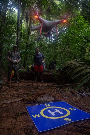 Dron blanco con luces rojas en el aire. Hombre con control y mujer en pie. Tapete azul de aterrizaje con 'H' y puntos cardinales en el suelo de tierra. Selva densa