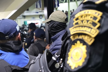 Vista trasera de varios agentes de policía con uniformes oscuros, gorras y un parche amarillo que dice 'COMISARIA 12', un casco de seguridad en primer plano