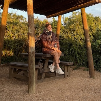 Mujer sentada en una mesa de pícnic de madera bajo una pérgola, sostiene una botella de agua. Alrededor hay vegetación verde y suelo de gravilla