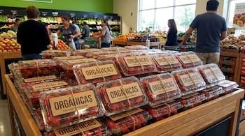 Múltiples envases transparentes de fresas orgánicas apilados en un expositor de madera en un supermercado. Se ven clientes y otras frutas y verduras al fondo.
