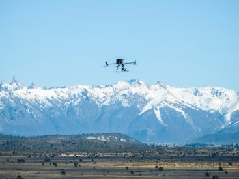 Un dron negro con cuatro hélices y una cámara vuela en el cielo azul. Al fondo, un paisaje de campo seco, árboles y montañas nevadas