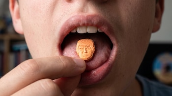 Primer plano de una boca abierta que recibe una pastilla naranja con la forma de la cara de Donald Trump.