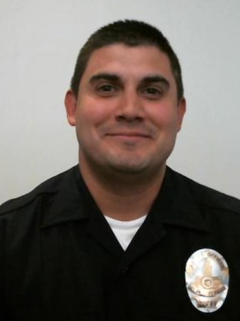 Retrato de un hombre sonriente con uniforme policial negro, camiseta blanca y una placa metálica dorada en el pecho sobre fondo blanco