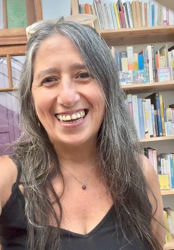 Retrato de Daniela Azulay, una mujer sonriente de cabello gris largo, camiseta negra y collar con dije oscuro. Al fondo, estanterías con libros