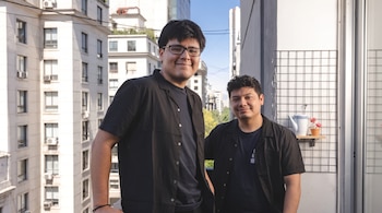 Dos hombres jóvenes con camisa negra posan en una terraza con grandes edificios residenciales de fondo.
