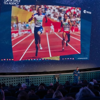 Una pantalla gigante muestra dos atletas con prótesis corriendo en una pista roja. Abajo, un astronauta da una charla a una audiencia sentada