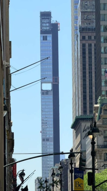 Rascacielos 262 Fifth Avenue en construcción, esbelto, con fachada de cristal y secciones oscuras, contra cielo azul. Se aprecian andamios y edificios de Manhattan