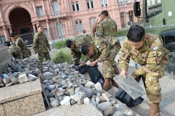 Cinco soldados con uniforme camuflado manipulan piedras y bolsas negras en el suelo, con la Casa Rosada y un camión militar en el fondo