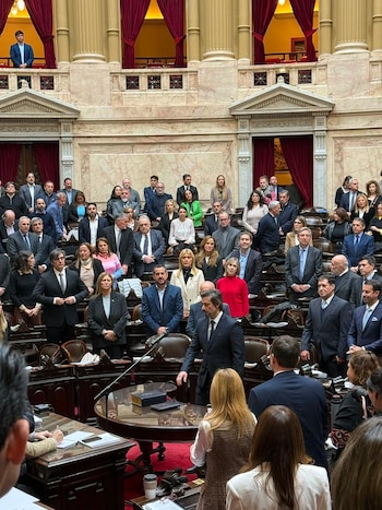 Una sala de sesiones con múltiples asientos de madera, alfombra roja, columnas y balcones. Numerosos hombres y mujeres de pie en el recinto