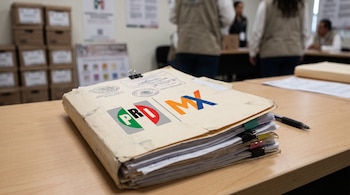 Primer plano de un folder con papeles, sellos, logo del PRI y logo de Somos México; un bolígrafo sobre una mesa de madera; fondo borroso con cajas y personas.