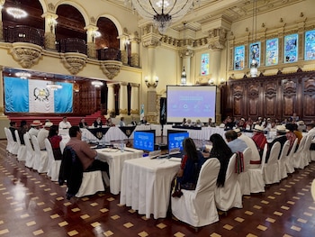 Interior de un gran salón adornado con columnas y balcones. Personas sentadas en sillas con fundas blancas alrededor de mesas. Una pantalla proyecta "Política Pública de Turismo Sostenible"