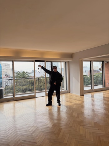 Hombre de ropa oscura y gafas de sol levanta un manojo de llaves en una sala vacía con piso de parquet, ventanales y balcón con árboles