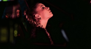 Wanna, cantante colombiana, de perfil, mirando hacia arriba, con una trenza lateral y un pendiente, bajo luz roja. Un asiento de coche con logo Lamborghini es visible