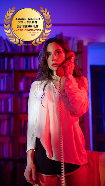 Una mujer de cabello castaño y blusa blanca sostiene un teléfono fijo rojo en su mejilla. La escena está iluminada con tonos rojos y azules