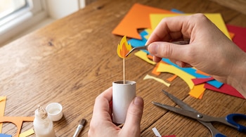 Unas manos utilizan pinzas para colocar una llama de cartulina en un tubo de cartón sobre una mesa con tijeras, pegamento y recortes de papel.